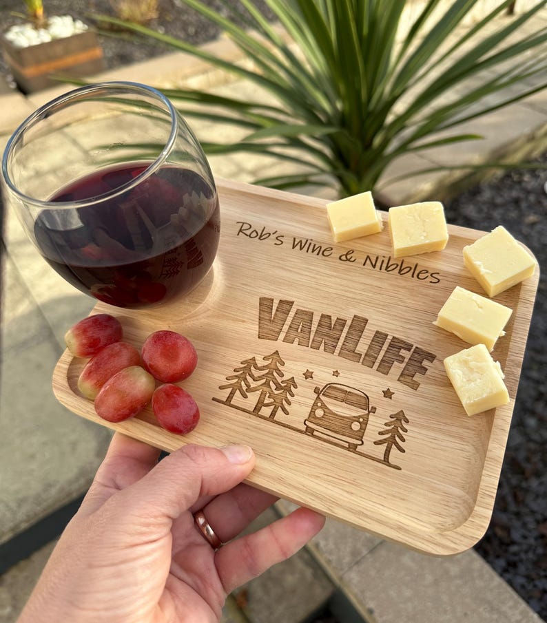 May include: A rectangular wooden serving tray with a circular cutout for a glass, engraved with the text "Rob's Wine & Nibbles" and "VANLIFE" above a graphic of a camper van and pine trees. The tray holds a glass of red wine, grapes, and cheese.