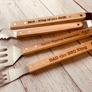 May include: Set of four grilling tools with wooden handles. The handles are engraved with the text "Rick - King of the Grill", "You're flipping awesome", "DAD the BBQ King", and a crossed grill and spatula symbol.
