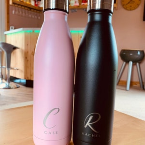 May include: Two insulated water bottles. One is pale pink with the name "Cass" and a cursive "C" engraved. The other is black with the name "Rachel" and a cursive "R" engraved. Both bottles have silver lids.