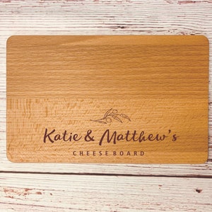 May include: A wooden cheese board with the text "Katie & Matthew's Cheese Board" engraved on it. The board has a light brown color and a natural wood grain.