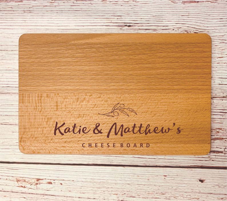May include: A wooden cheese board with the text "Katie & Matthew's Cheese Board" engraved on it. The board has a light brown color and a natural wood grain.