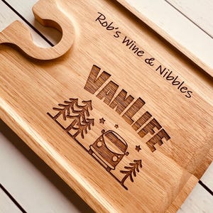 May include: A rectangular light-coloured wooden serving board featuring a cutout for a wine glass stem. The board is engraved with the text "Rob's Wine & Nibbles" and "VANLIFE" alongside a graphic of a camper van, pine trees, and small stars.