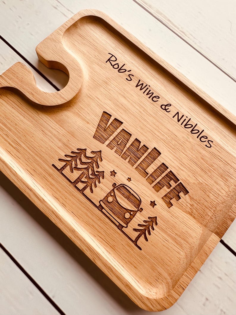 May include: A rectangular light-coloured wooden serving board featuring a cutout for a wine glass stem. The board is engraved with the text "Rob's Wine & Nibbles" and "VANLIFE" alongside a graphic of a camper van, pine trees, and small stars.