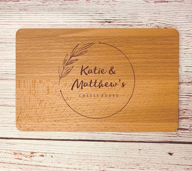 May include: A wooden cheese board with a circle design engraved with the text "Katie & Matthew's Cheese Board" and a leafy branch design.