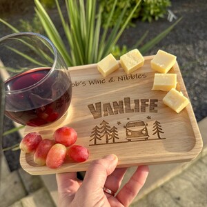 May include: A rectangular light-coloured wooden serving tray featuring the engraved text "Bob's Wine & Nibbles" and "VANLIFE" with a camper van and pine tree graphic. The tray holds a glass of red wine, several red grapes, and cubes of cheese.