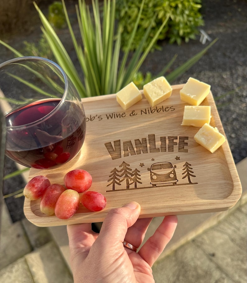 May include: A rectangular light-coloured wooden serving tray featuring the engraved text "Bob's Wine & Nibbles" and "VANLIFE" with a camper van and pine tree graphic. The tray holds a glass of red wine, several red grapes, and cubes of cheese.