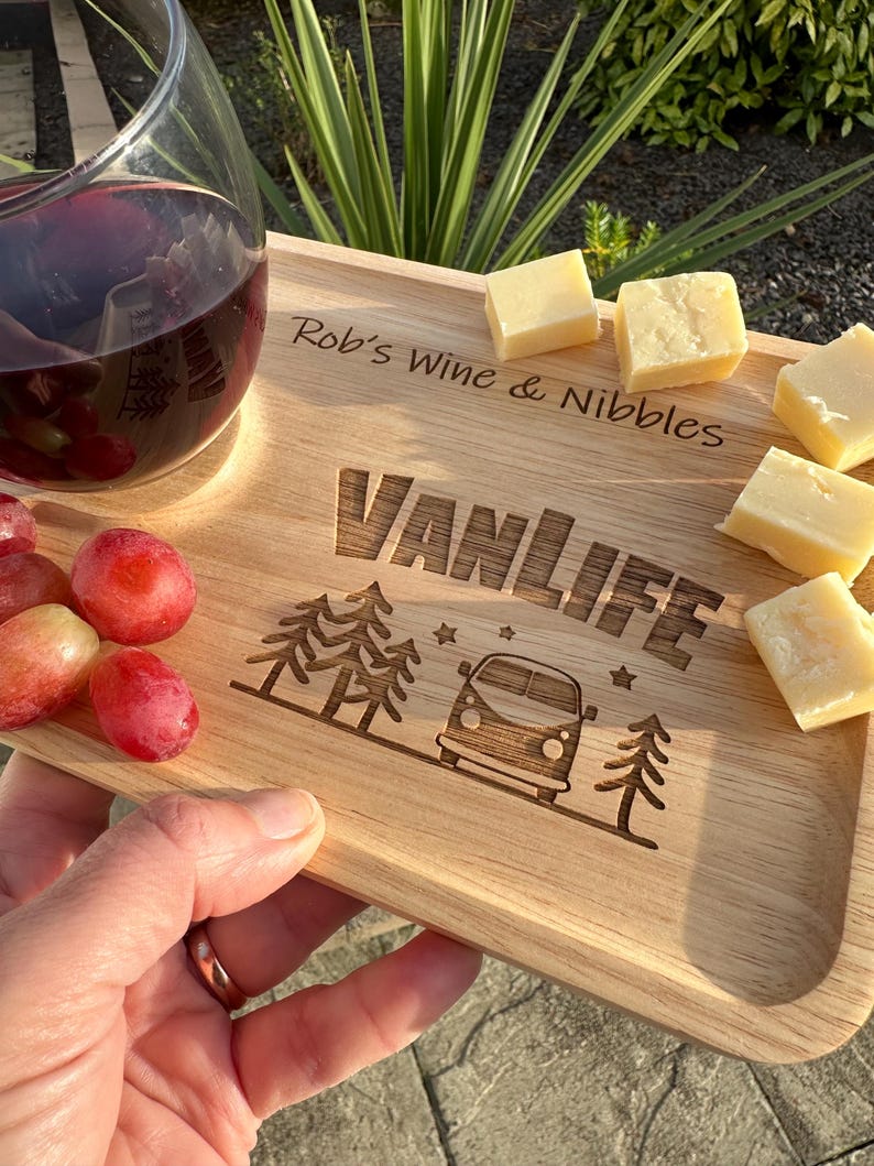 May include: A rectangular wooden serving board featuring the engraved text "Rob's Wine & Nibbles" and "VANLIFE" alongside a graphic of a camper van and pine trees. The board holds a glass of red wine, several red grapes, and cubes of cheese.