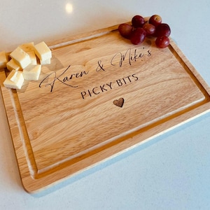 May include: A rectangular wooden serving board with a carved border. The board is engraved with the text "Karen & Mike's PICKY BITS" and a heart symbol. Cubes of cheese and red grapes are arranged on the board.