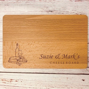 May include: A wooden cheese board with an engraved design of a wine bottle, glass, cheese, and grapes. The text "Suzie & Mark's Cheese Board" is engraved below the design.