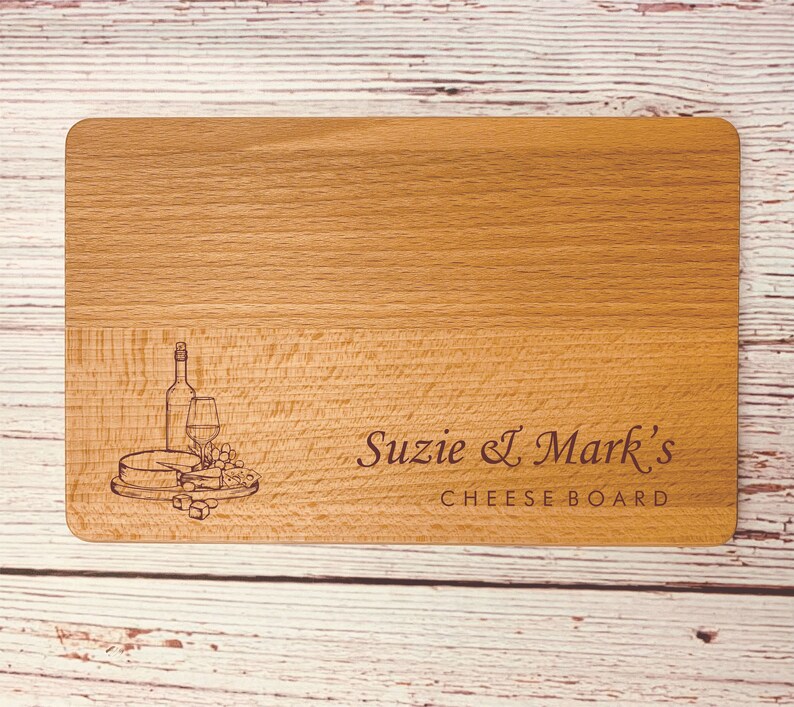 May include: A wooden cheese board with an engraved design of a wine bottle, glass, cheese, and grapes. The text "Suzie & Mark's Cheese Board" is engraved below the design.