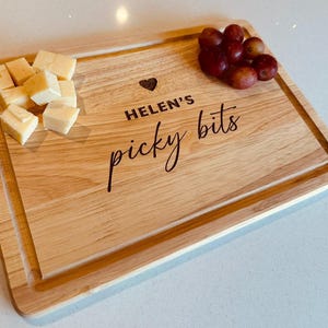 May include: A wooden serving board with the text "HELEN'S picky bits" and a heart symbol. It is filled with cheese cubes and red grapes. The board has a recessed edge, perfect for serving snacks.