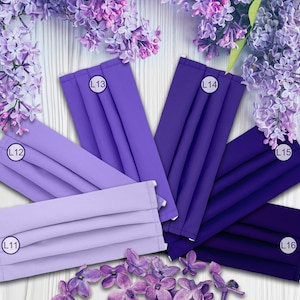 May include: Six different shades of purple fabric face masks, labeled L11, L12, L13, L14, L15, and L16. The masks are arranged in a semi-circle on a white wooden surface with purple flowers.