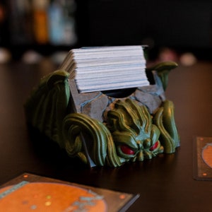 May include: A green and brown plastic card holder shaped like a monster with red eyes. The holder is filled with a stack of white cards. Three orange and blue cards are on the table in front of the holder.