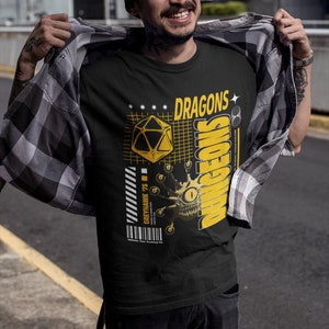 Op de afbeelding: Een zwart T-shirt met een geel grafisch ontwerp met een Dungeons and Dragons logo, een 20-zijdige dobbelsteen en een drakenoog. De tekst "DRAGONS & DUNGEONS" is geschreven in een gestileerd lettertype.