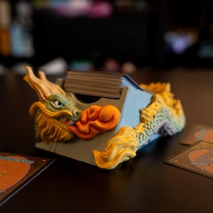May include: A blue, green, and orange dragon-shaped card holder with a stack of cards inside. The holder is on a dark brown wooden surface with three playing cards lying on the surface.