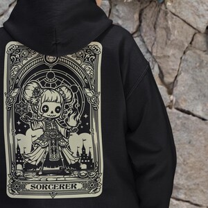 May include: Black hoodie featuring a white graphic print of a cartoon character dressed as a sorcerer. The graphic is framed in an ornate border and includes the word "SORCERER".
