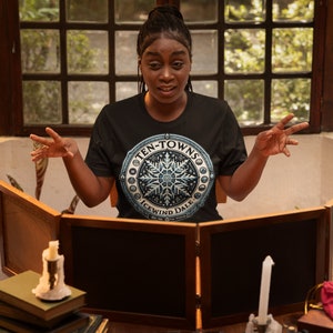 May include: A person wearing a black t-shirt with a blue and white snowflake design. The shirt says "Ten-Towns Icewind Dale". The person is sitting at a table with a wooden game board in front of them.