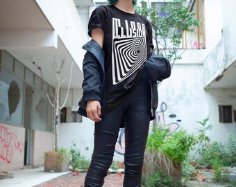 May include: A person wearing a black t-shirt with a white and black spiral design and the word "CLAR" in white block letters. The person is wearing black pants, black shoes, and a dark blue jacket. The background is an urban setting with brick and concrete.