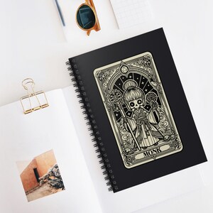 Puede incluir: Un cuaderno negro con espiral con una pegatina de carta de tarot color crema que representa a un monje esqueleto sosteniendo un bastón. La pegatina tiene intrincados dibujos lineales negros y la palabra "MONK". El cuaderno está abierto sobre una superficie blanca.