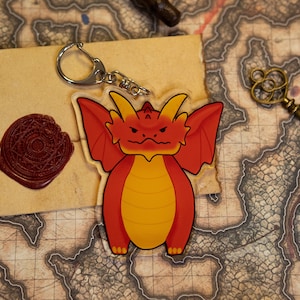 May include: Red and yellow dragon keychain with a silver key ring. The dragon has a grumpy expression and is standing on its hind legs.