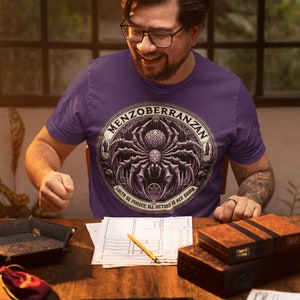 Może przedstawiać: Osoba w fioletowym t-shircie z grafiką pająka i tekstem "Menzoberranzan.  Loth be praised, all victory is her doing."