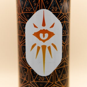 May include: A white tumbler with a black and orange design featuring a repeating pattern of 20-sided dice. A white sticker with an orange eye and radiating lines is centered on the tumbler.
