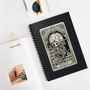 Puede incluir: Un cuaderno espiral en blanco y negro con una ilustración de un personaje pícaro. La ilustración está en el estilo de una carta de tarot con un fondo negro y una carta de color crema con detalles ornamentados. La carta dice "Rogue".