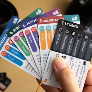 Op de afbeelding: Een hand houdt een waaier van fotografie cheat sheets met verschillende instellingen en iconen. De kaarten zijn gelabeld met termen als "LANDSCHAP", "VOGEL" en "MAAN". Een cameralens en camerabody zijn zichtbaar op de achtergrond.