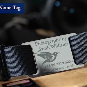 May include: A silver camera name tag with the text "Photography by Sarah Williams" and a bird design. The tag also includes a phone number and email address: +44 20 7219 3000, swilliams32@email.co.uk.