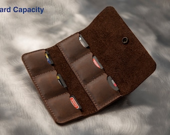 Könnte beinhalten: Ein brauner Lederkartenhalter mit sechs Steckplätzen für Spielkarten. Der Halter ist geöffnet und die Steckplätze sind mit bunten Spielkarten gefüllt. Der Text "6 Card Capacity" ist in der oberen linken Ecke des Bildes sichtbar.