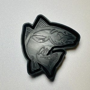 May include: A black, 3D-printed fish-shaped object with detailed lines and a textured surface. The fish is outlined in black, with fins and scales visible. The design is likely for crafting or decoration.