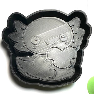 Könnte beinhalten: Schwarzer Plastik-Ausstecher in Form eines Axolotls, der ein Herz hält. Der Axolotl hat große Augen und ein kleines Lächeln.