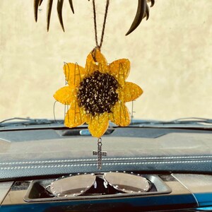 May include: A yellow sunflower car air freshener with a rhinestone cross hanging from it. The air freshener is hanging from a rearview mirror in a car.