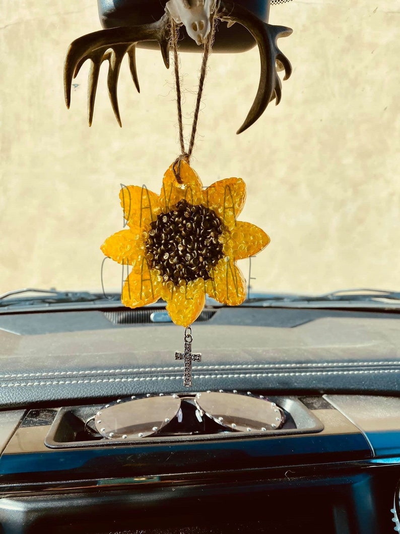 May include: A yellow sunflower car air freshener with a rhinestone cross hanging from it. The air freshener is hanging from a rearview mirror in a car.