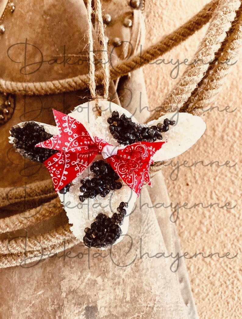 May include: White cow ornament with black spots and a red bandana. The ornament is hanging from a rope.