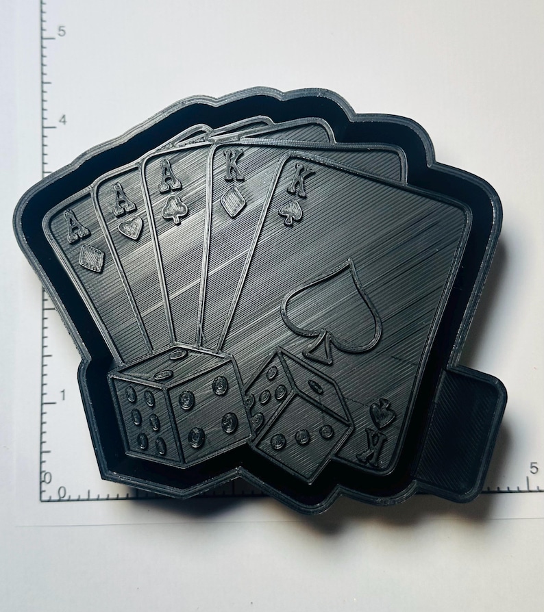 May include: Black cookie cutter featuring a poker hand of playing cards: Ace of Diamonds, Ace of Spades, Ace of Hearts, Ace of Clubs, King of Diamonds, King of Spades, and two dice. The cookie cutter is approximately 5 inches wide.