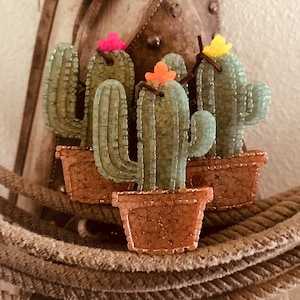 May include: Four small cactus ornaments with green bodies, brown pots, and colorful flower accents. The ornaments are arranged on a brown rope background.