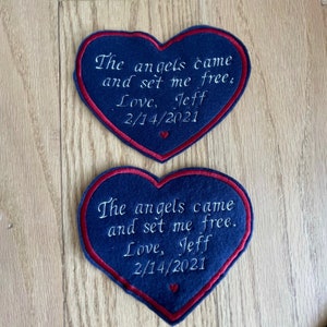 Może przedstawiać: Dwa sercowate łaty z niebieskiego filcu z czerwonymi szwami. Każda łata ma wyszyty białymi nićmi tekst "The angels came and set me free. Love, Jeff 2/14/2021".