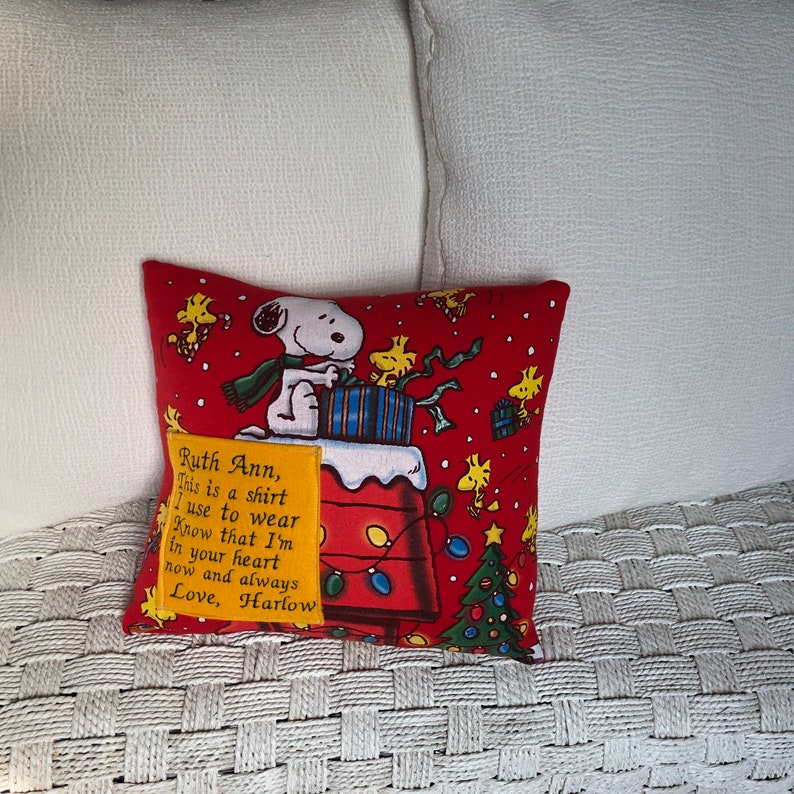 Może przedstawiać: Czerwono-ż&oacute;łta poduszka z motywem świątecznym Snoopy'ego. Poduszka ma ż&oacute;łtą łatę z tekstem "Ruth Ann, This is a shirt I use to wear Know that I'm in your heart now and always Love, Harlow".