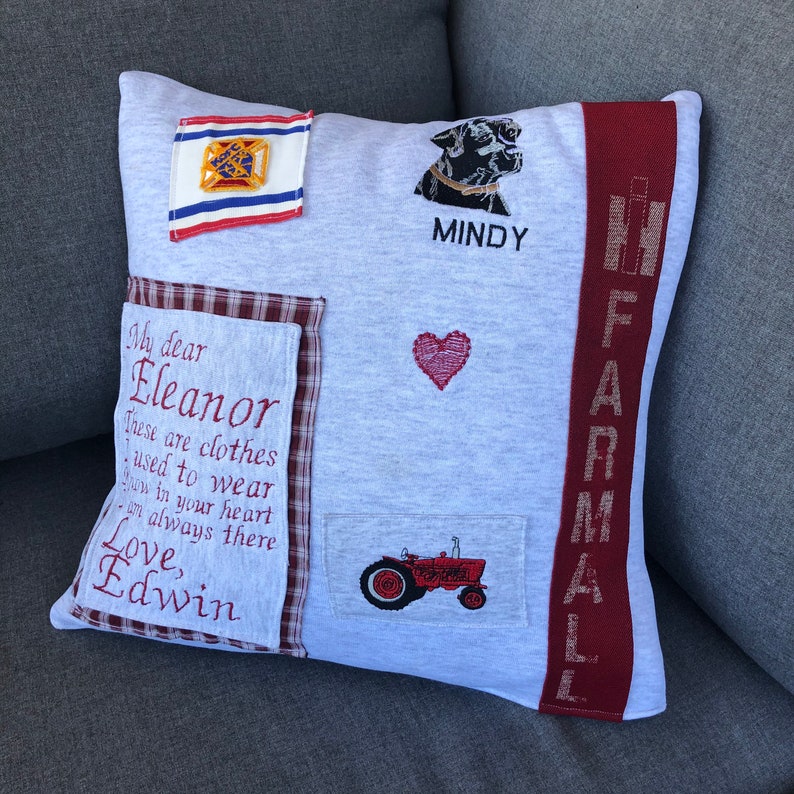 Może przedstawiać: Szara poduszka z czerwonymi, białymi i niebieskimi łatami. Poduszka ma wyszyte napisy: "My dear Eleanor These are clothes used to wear now in your heart I'm always there Love, Edwin" oraz "MINDY" i "FARMALL". Na poduszce wyszyto r&oacute;wnież czerwone serce, czarnego psa i czerwony traktor.
