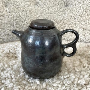 Könnte beinhalten: Kleine, dunkel glasierte Keramik-Teekanne mit rundem Körper, gebogenem Henkel und kleinem Ausguss. Der flache Deckel hat die gleiche Farbe wie die Teekanne. Die Teekanne hat eine dunkle, gesprenkelte Farbe mit Andeutungen von Blau und Grün.