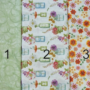 May include: Three fabric swatches, each with a different pattern. The first swatch is a light green with a swirl pattern. The second swatch is white with a floral pattern and jars. The third swatch is white with a floral pattern in shades of orange, yellow, and pink.