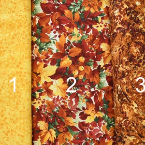 May include: Three fabric swatches, each with a different pattern. The first swatch is a solid yellow with a subtle pattern of small white dots. The second swatch features a colourful autumn leaf pattern with red, orange, yellow, and green leaves. The third swatch is a brown and orange abstract pattern.