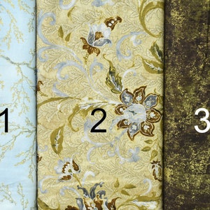 May include: Three fabric swatches, each with a different pattern. The first swatch is light blue with a white floral pattern. The second swatch is a cream color with a gold and silver floral pattern. The third swatch is a dark green with a gold and brown speckled pattern.