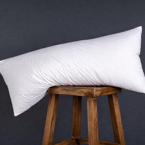 Puede incluir: Una almohada larga y blanca para el cuerpo descansa sobre un pequeño taburete de madera. La almohada está en ángulo, con una apariencia suave y esponjosa. El taburete tiene un acabado de madera natural y está sobre un fondo gris oscuro.