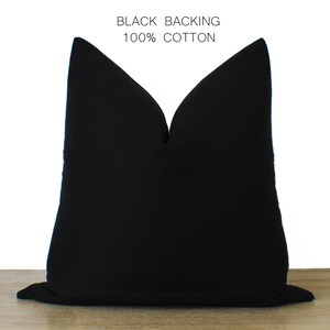 May include: A solid black pillow with the text "BLACK BACKING 100% COTTON" in white. The pillow is a square shape and appears to be made of a soft fabric. The pillow is sitting on a wooden surface.