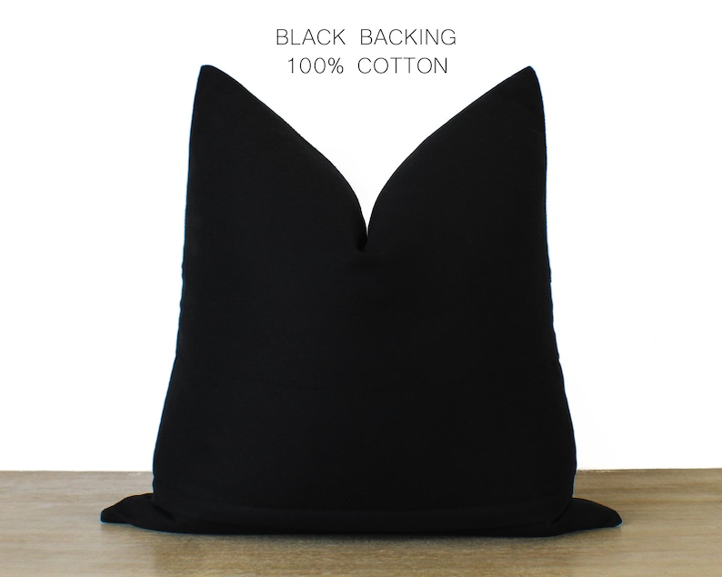 May include: A solid black pillow with the text "BLACK BACKING 100% COTTON" in white. The pillow is a square shape and appears to be made of a soft fabric. The pillow is sitting on a wooden surface.