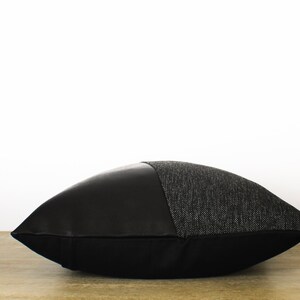 May include: A decorative pillow with a two-tone design. The top half is a textured gray fabric, and the bottom half is smooth black. The pillow is resting on a wooden surface against a white background.