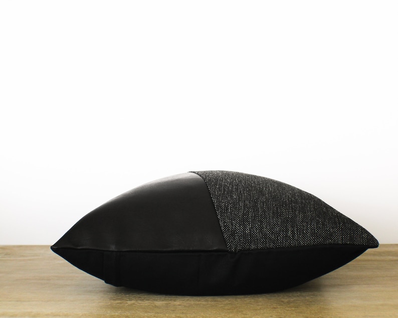 May include: A decorative pillow with a two-tone design. The top half is a textured gray fabric, and the bottom half is smooth black. The pillow is resting on a wooden surface against a white background.
