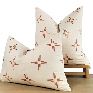 May include: Two decorative throw pillows with a white background and a rust-colored geometric pattern. The pillows are on a wooden surface.